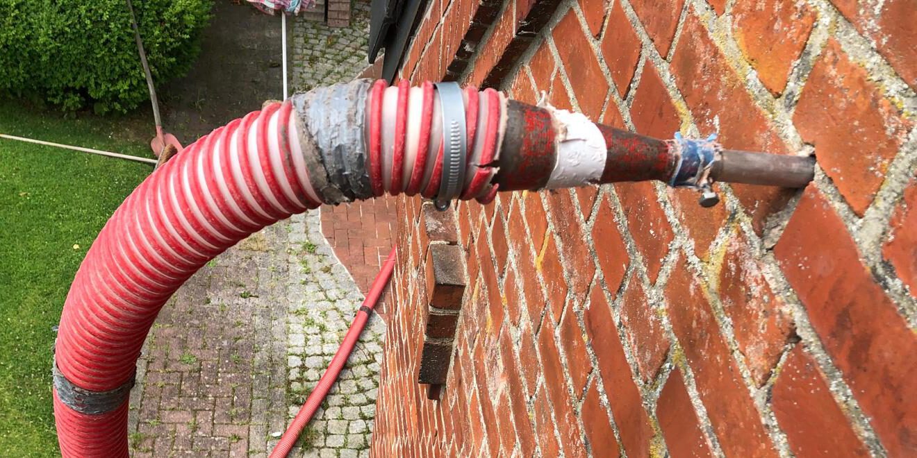 Roter Wellschlauch, der mit einer Schlauchschelle an ein Metallrohr angeschlossen ist, das durch eine Ziegelwand führt